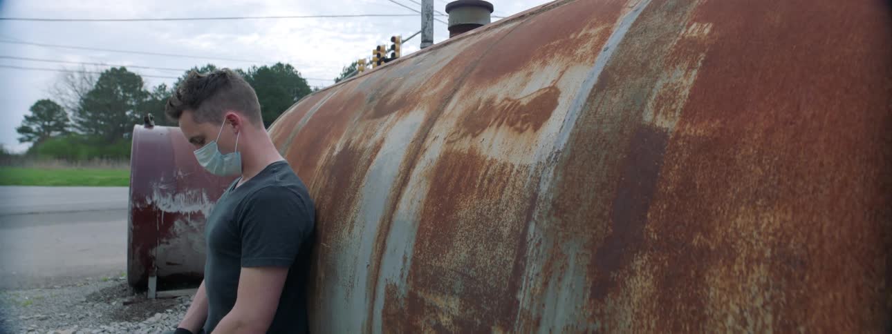 A teenage boy leans against an oil tank wearing a medical style face mask