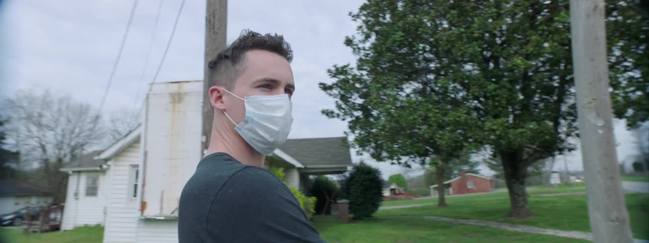 A man wearing a protective face mask stands outdoors in a small rural community