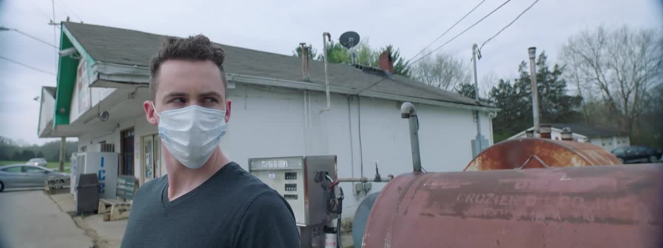 A white man stands outdoors wearing a face mask and looking off into the distance