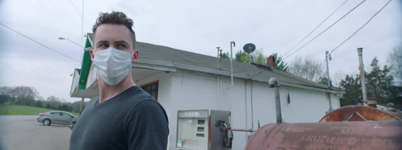 A white man wearing a face mask stands outside a gas station and looks around