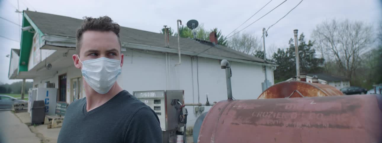A white man wearing a face mask stands near a gas station and takes in his surroundings
