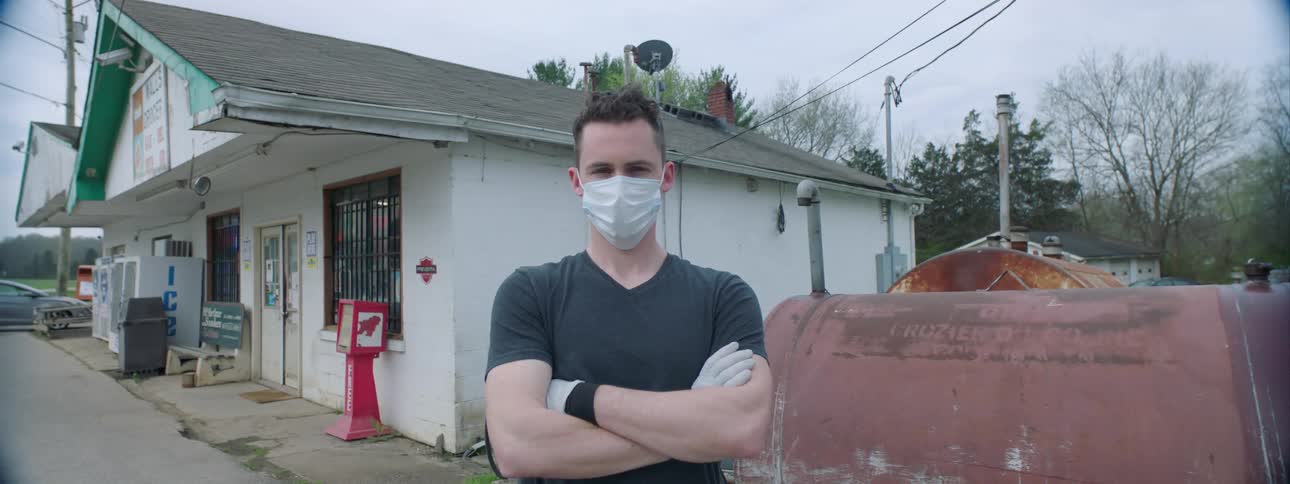 A man stands in front of a convenience store wearing gloves and a medical mask