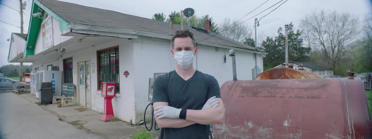 A man in a face mask and latex gloves sitting outside a small store while crossing his arms