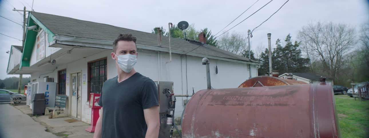 A man wearing a protective mask stands near a rusted tank and gas pump at a rural station