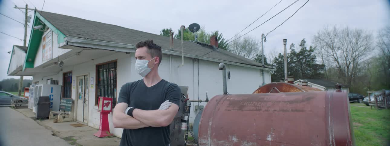 A man wearing a protective mask stands with arms crossed next to a rusted tank and gas pump at a gas station