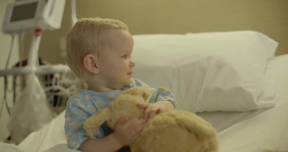 Blonde toddler child sitting on the bed is being given a doll and he plays with the doll in the hospital