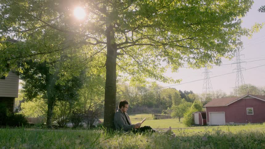 A man sits at the base of a tree and reads a book