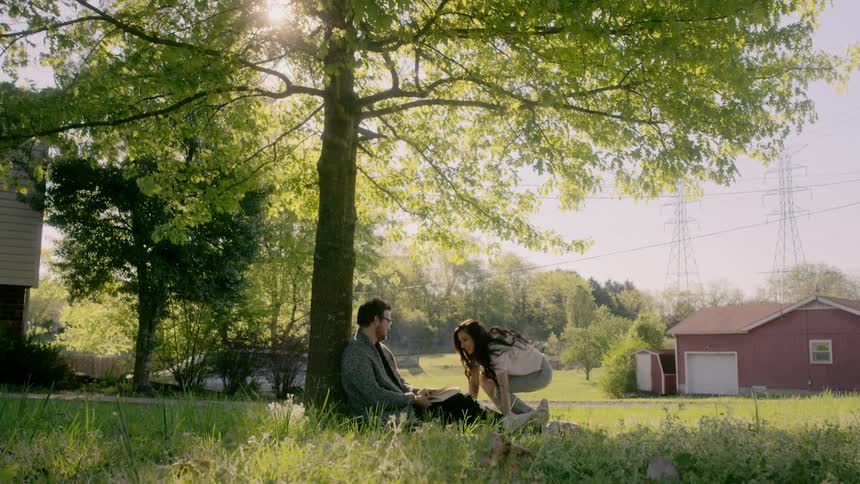 A woman coming to sit next to a man who is sitting against a tree reading a book