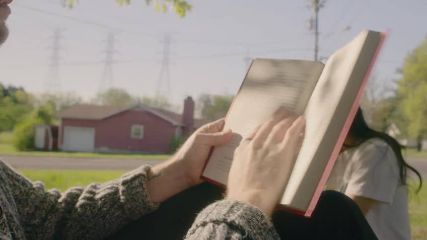 A man reads a book as a woman sits on the grass by him
