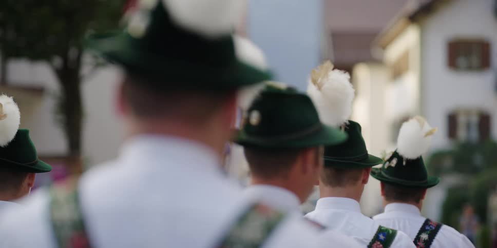Men wearing lederhosen walk down the middle of a village street playing drums