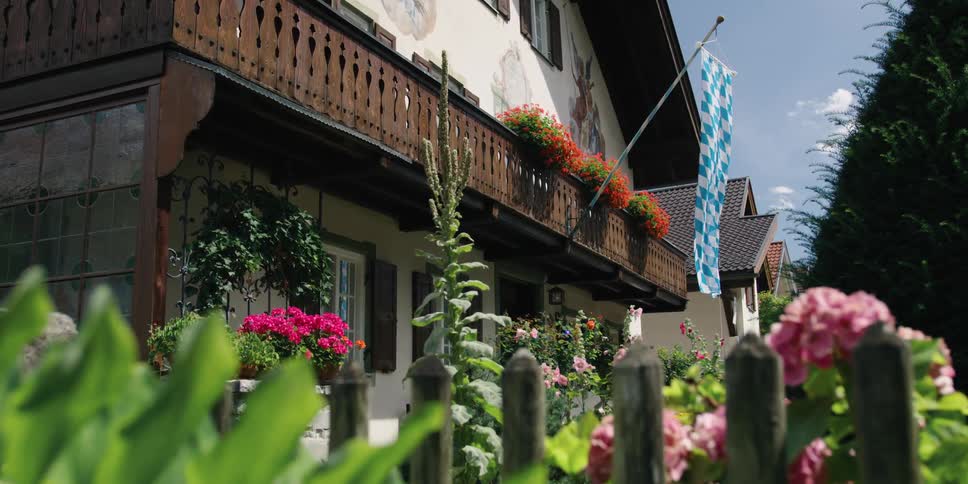 A blue and white checkered banner hanging from a flag pole on a balcony sways in the breeze over a colorful garden