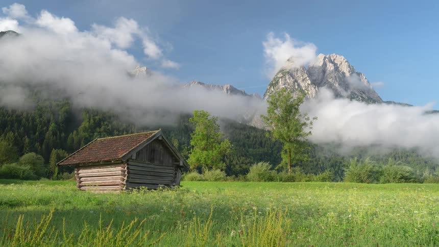 A shack sits in a field as clouds surround the mountains