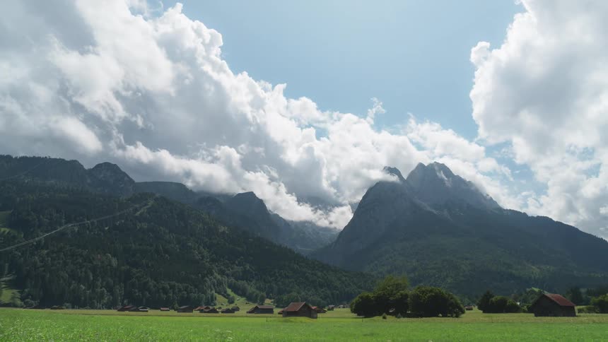 White clouds in a blue sky pass over mountain peaks and green grass with cabins