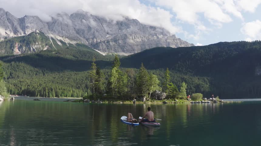 A man straddling his paddle board on a lake holds onto his girlfriends board where she is lying down as they both float along