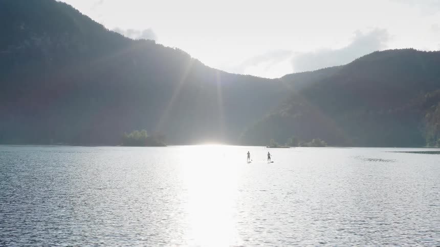 Three people stand and paddle on boards in lake near mountain under glaring sun
