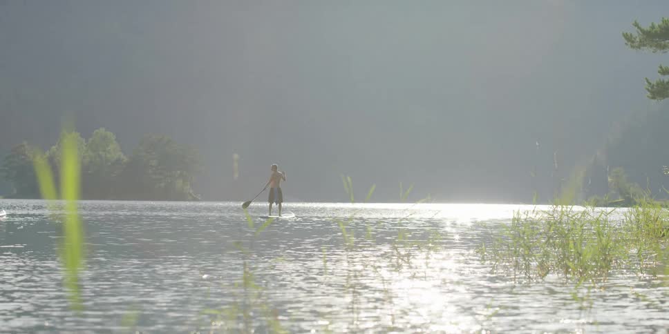 Two men stand up on surfboards paddling in river as light glares off the water