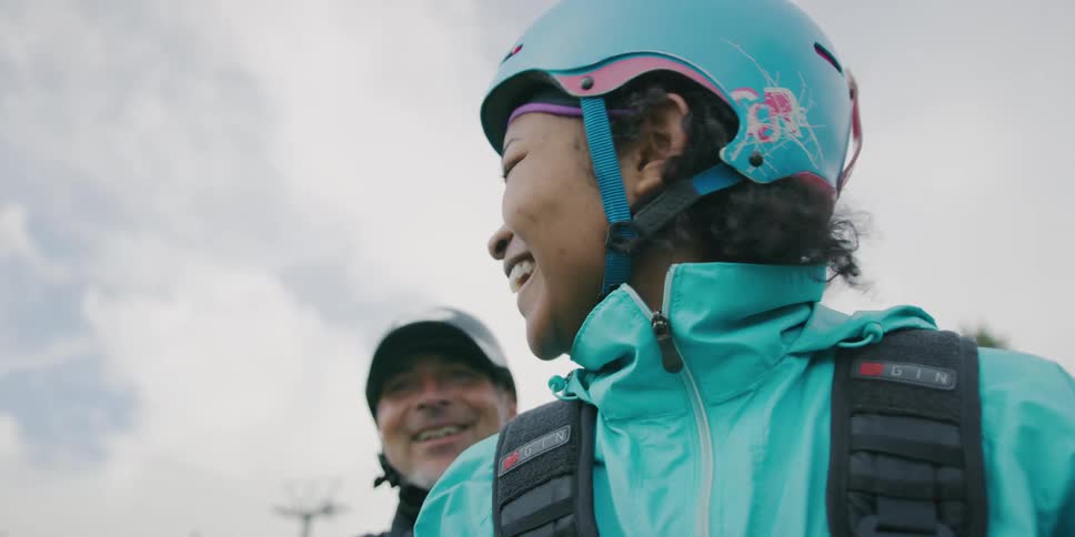 A woman wearing safety gear and a helmet laughs while talking to a man standing behind her