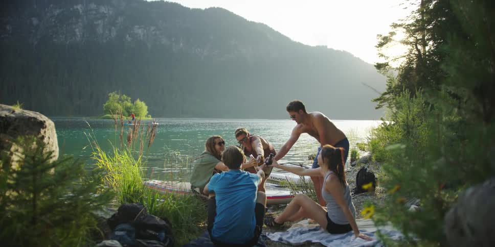 A group of people knocking beer bottles together on the shore of lake during the day