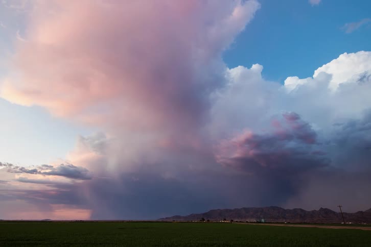 Storm clouds cover a blue sky and lightning flashes down over a green field and cars on a road