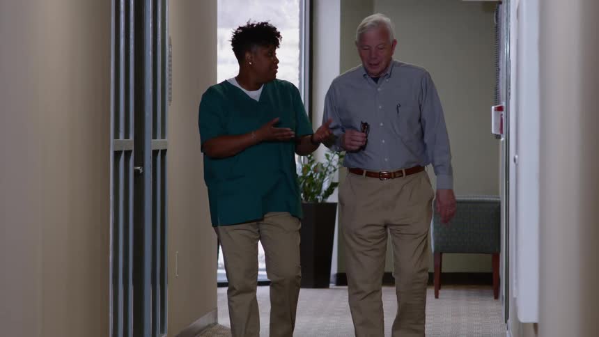 A nurse walks with an elderly male patient down the hallway of a clinic