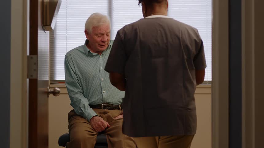 Elderly patient sitting on hospital bed as nurse walks in to inspect injury