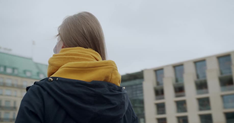 A teen wearing a mask stands outside buildings in a city