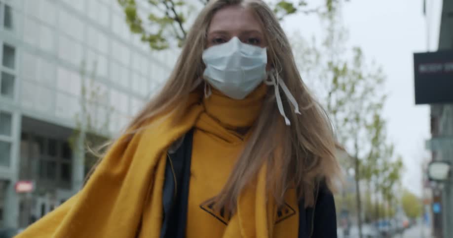 A teenager wearing a mask walks down a quiet city street and turns to look behind her