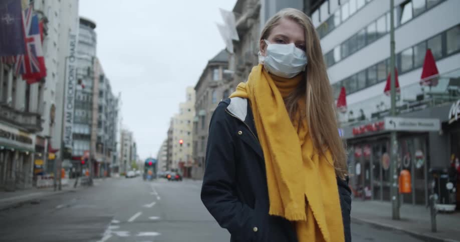 A woman wearing a mask stands on an empty city street with the wind blowing her hair and scarf