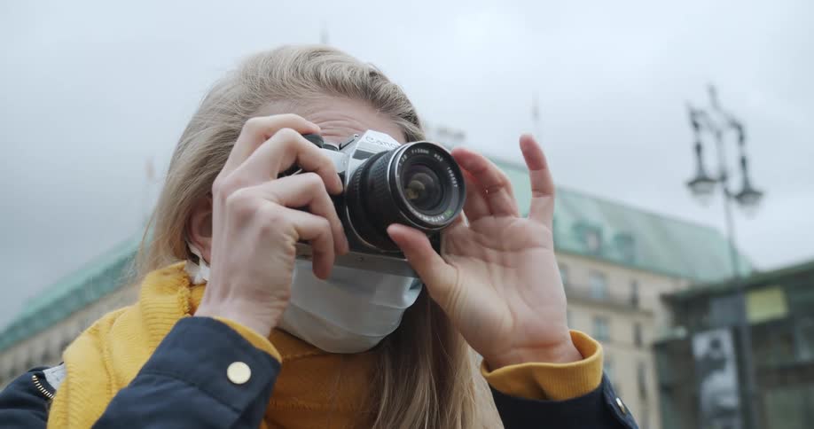A woman wearing a mask holds up and focuses a camera to take a photo in a city on a cloudy day