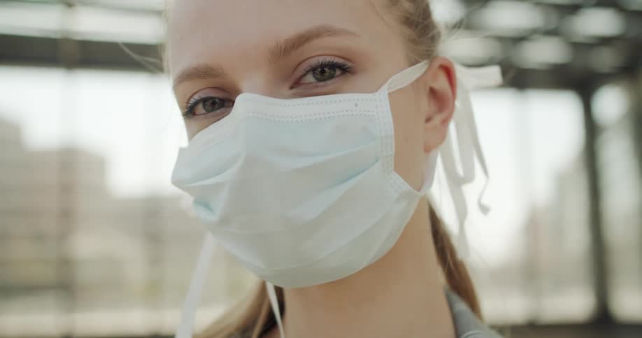Blonde woman wearing a white face mask due to coronavirus looks at the camera