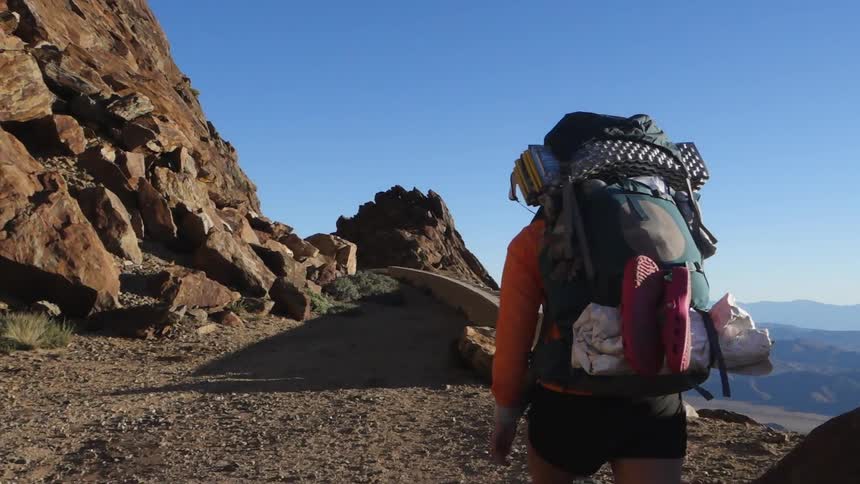A hiker, carrying a large backpack, travelling on a mountainside trail high above a valley with more mountains visible in the background