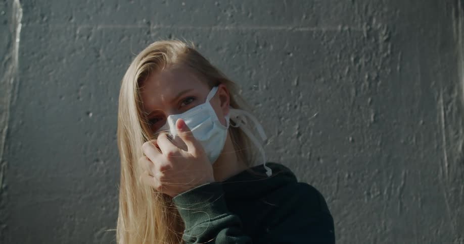 Blonde woman wearing a white face mask due to corona virus stands in front of the wall outdoors