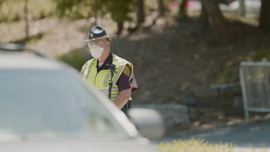 A state trooper stands with hands folded wearing a mask watching cars pass by