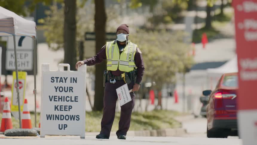 A police officer with a mask and a yellow vest on standing in the street with a sign in his hand