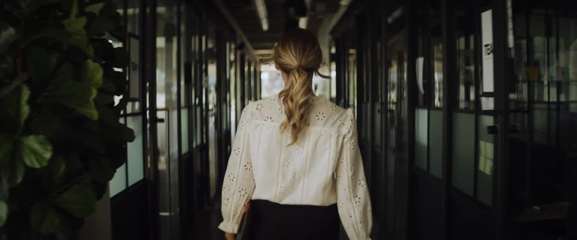 A woman carrying files walks down a hallway passing offices with her ponytail bouncing on her back