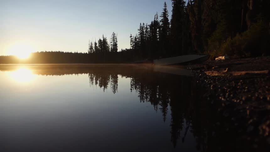 The sun setting behind a tree-line and reflecting off a lake as fog begins to rise near a beached boat