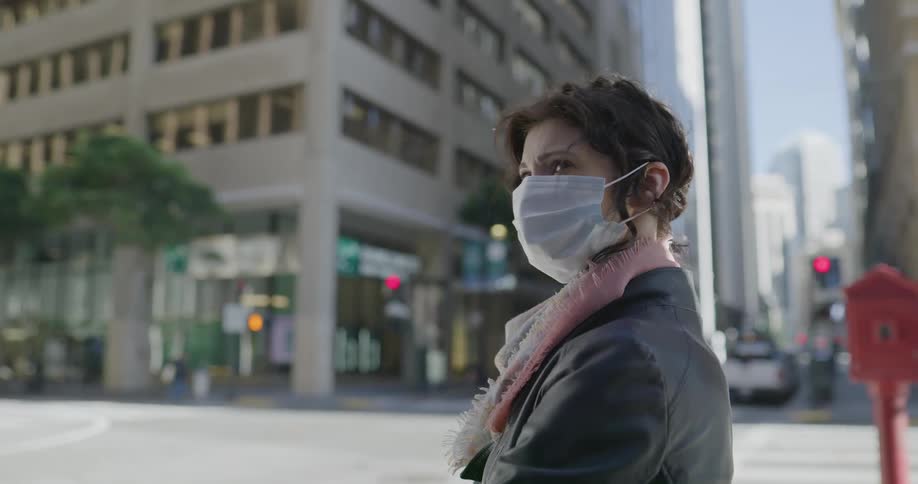 A woman stands at an intersection in a city wearing a mask