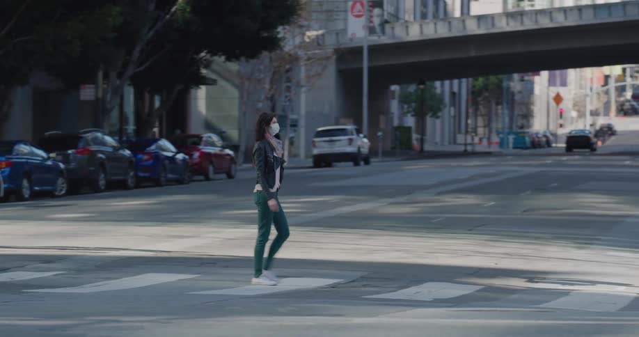 A woman wearing a surgical mask crosses a city street as vehicles pass by