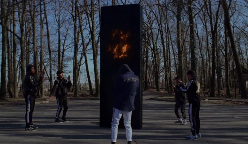 A group of men dance choreographed steps around a large black panel glowing orange in a parking lot
