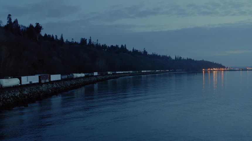 Slow pullback as a freight train travels beside calm water at twilight