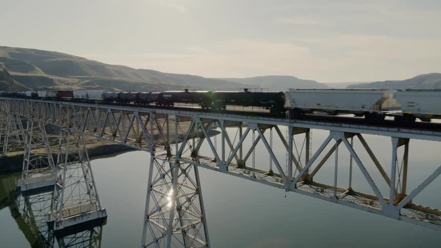Freight train rolling along a high trestle above glass calm water reflecting the sun