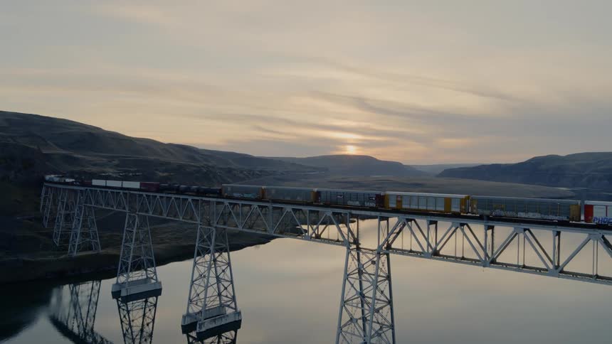 A freight train traveling across a long and tall bridge over water at sunset