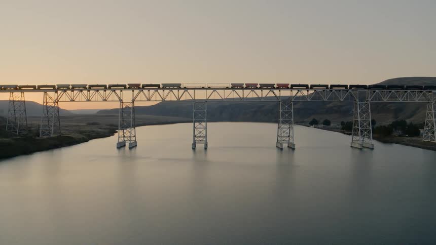 Push towards a freight train crossing a river on a long bridge as sky glows orange on the horizon