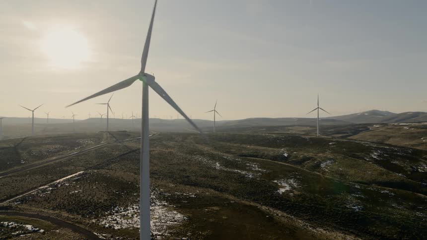 Revealing a wind turbine passing in front of the sun over snow dotted rolling hils