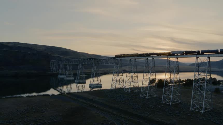 Flying parallel to a freight train starting across a tall bridge over mirror calm water