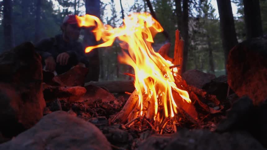 A camper sits at a campsite by a fire and opens a bag of food