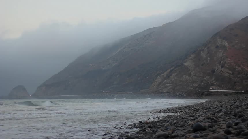 Choppy waves crash on a rocky shore near a mountain on a foggy day