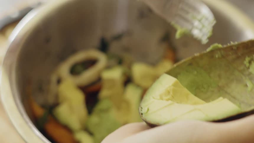 Woman scoops avocado out of shell and adds it to ingredients in a metal bowl