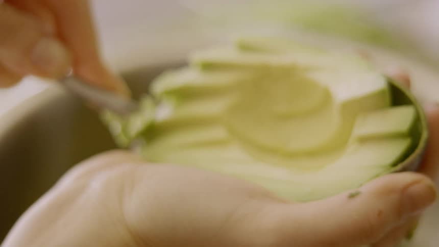A woman removes avocado with a spoon and adds it to a bowl of ingredients for guacamole