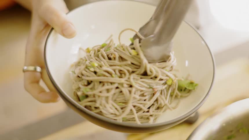 A person uses tongs to place noodles and vegetables into a bowl and artfully arrange it